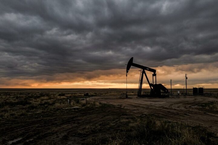 Pump jack silhouetted against a heavy overcast dusk, muted orange breaking through grey cloud, flat terrain running to...