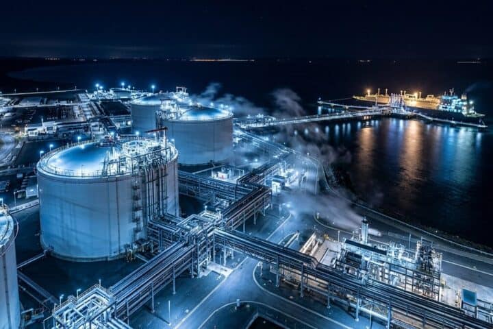 LNG export terminal at night: cryogenic storage tanks and insulated pipework lit against dark water, vapour drifting